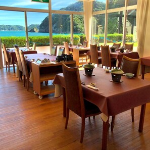 A restaurant with tables set for dining, large windows offering a view of the sea and hills, and a wooden floor. - Hotel Nagisaya (Nachikatsuura)
