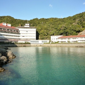 A waterfront hotel with multiple buildings, a clear view of the water, and a forested hillside in the background. - Hotel Nagisaya (Nachikatsuura)