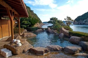 A traditional Japanese onsen with a wooden structure, large rocks, and a view of the sea. - Hotel Nagisaya (Nachikatsuura)