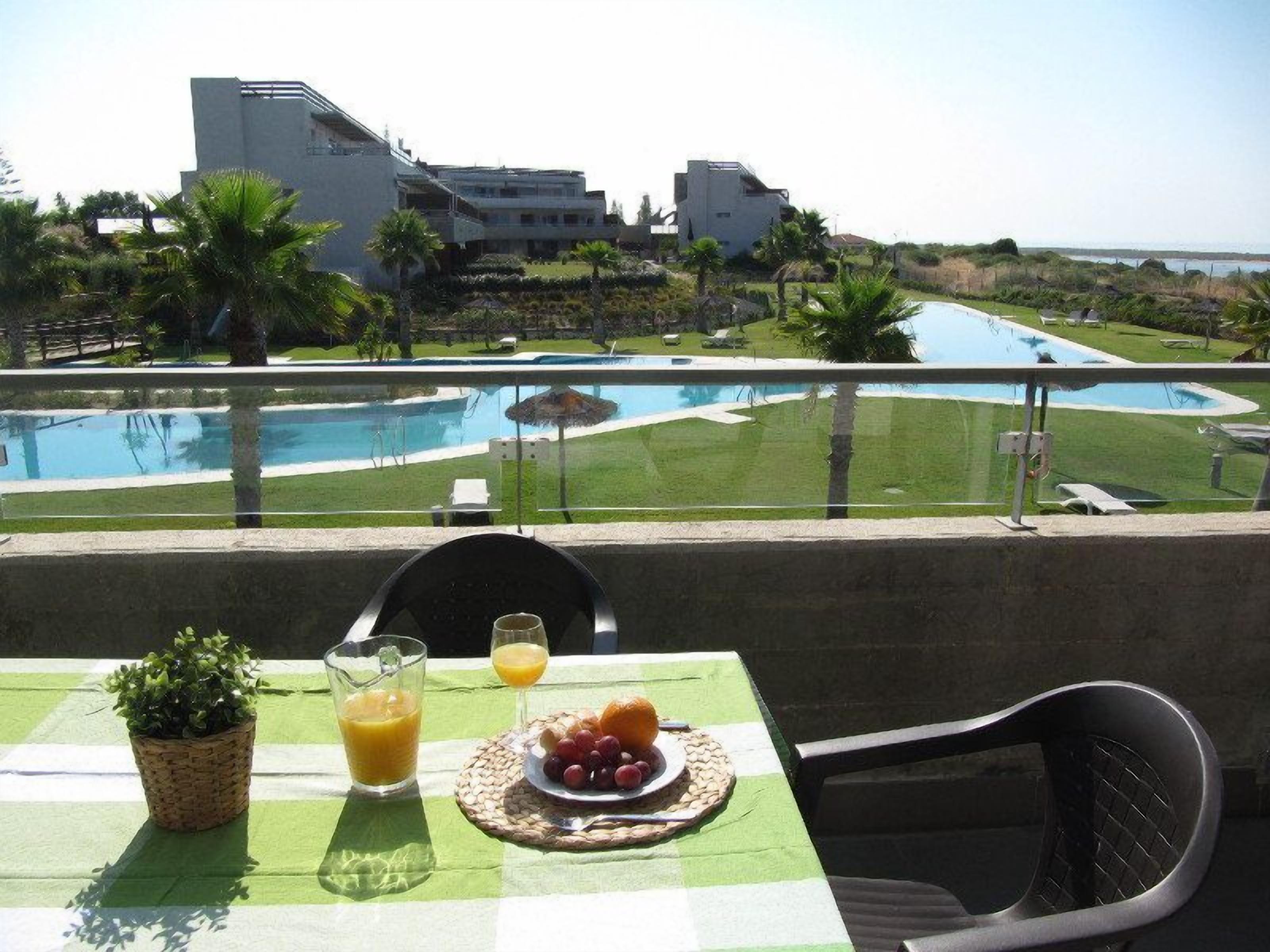 Un balcón con una mesa lista para el desayuno, con vista a una piscina y edificios con vegetación.