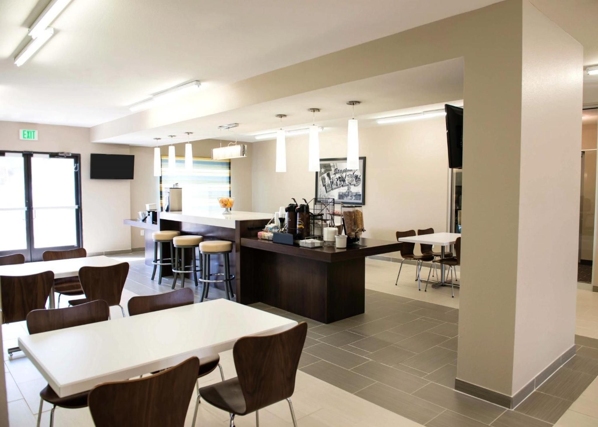 A modern cafeteria with high tables and chairs, a central serving area, and a flat-screen TV mounted on the wall.