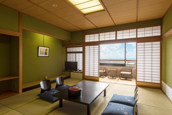 Una habitación japonesa tradicional con suelo de tatami, una mesa baja, sillas, un televisor y vistas a través de biombos shoji.