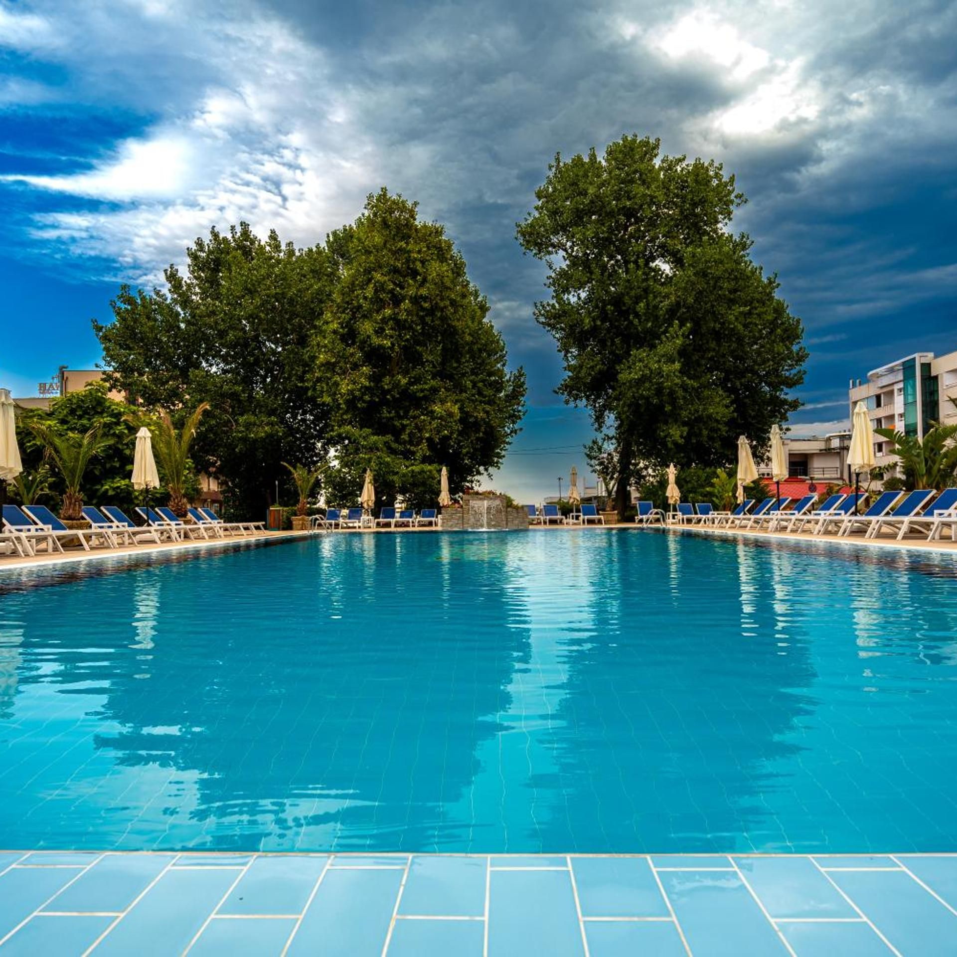 A swimming pool with lounge chairs and umbrellas, surrounded by trees and buildings.