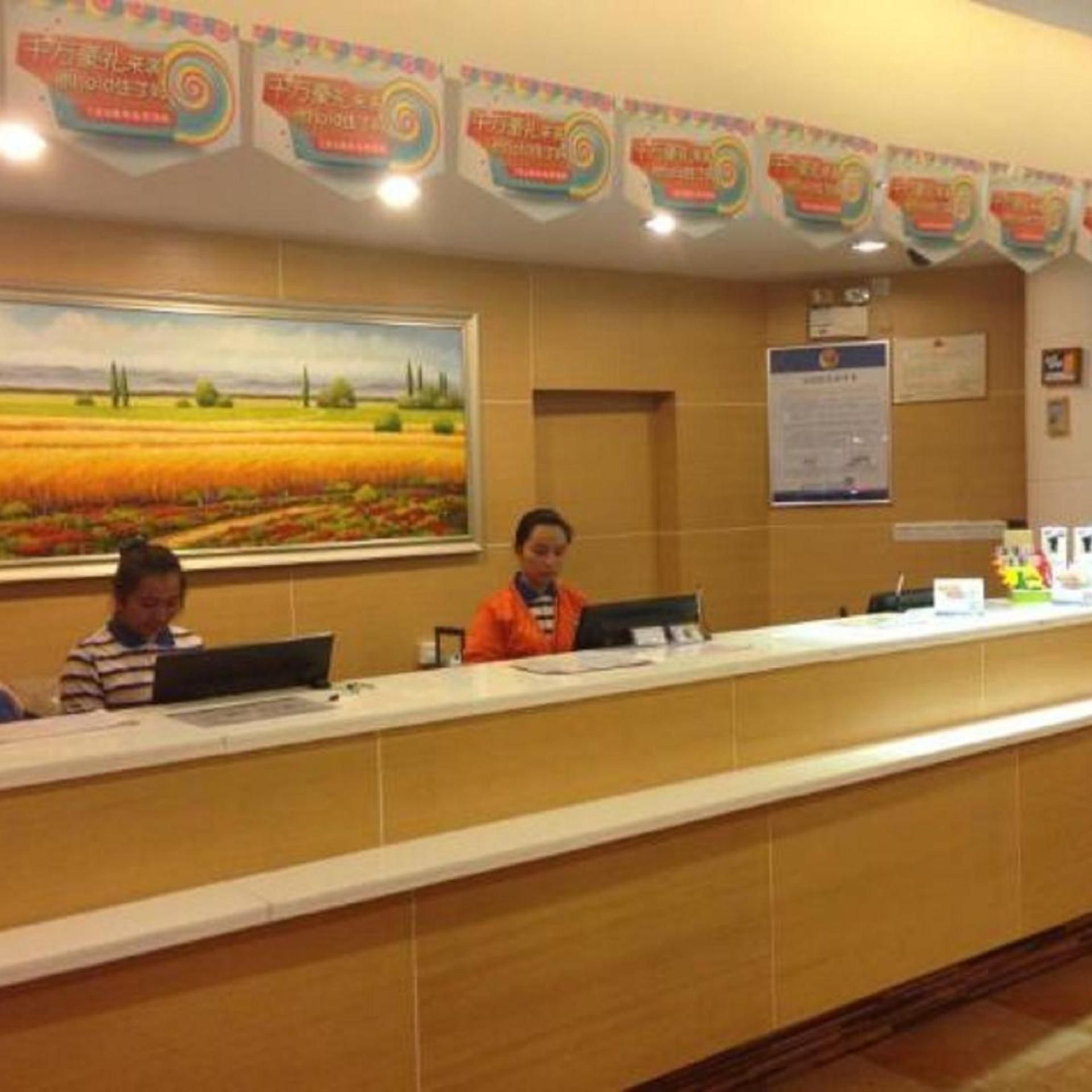 A reception desk with two staff members seated, a painting of a field on the wall, and a bulletin board with notices.