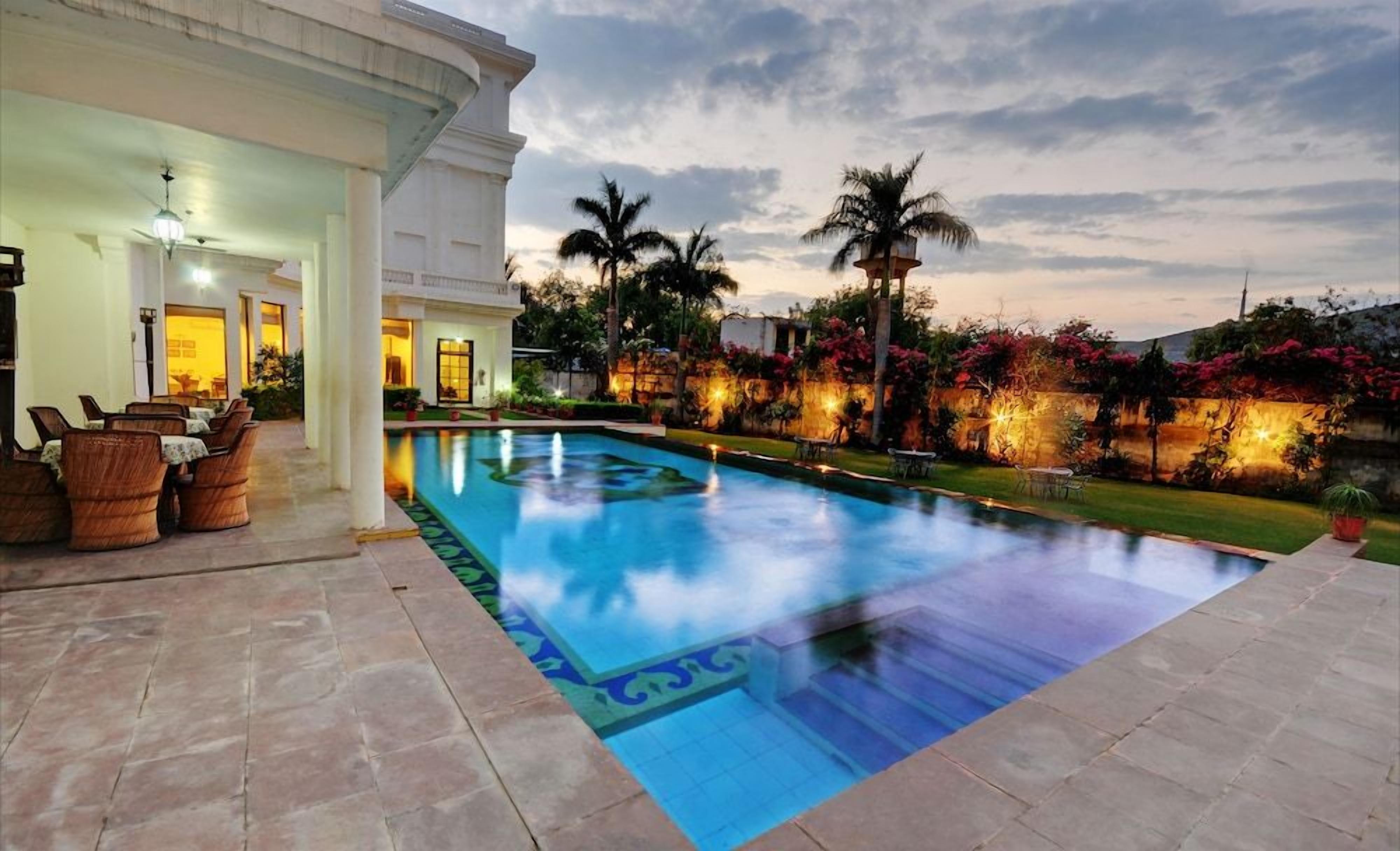 A swimming pool with a tiled deck, surrounded by palm trees and flowering plants, with a building in the background.