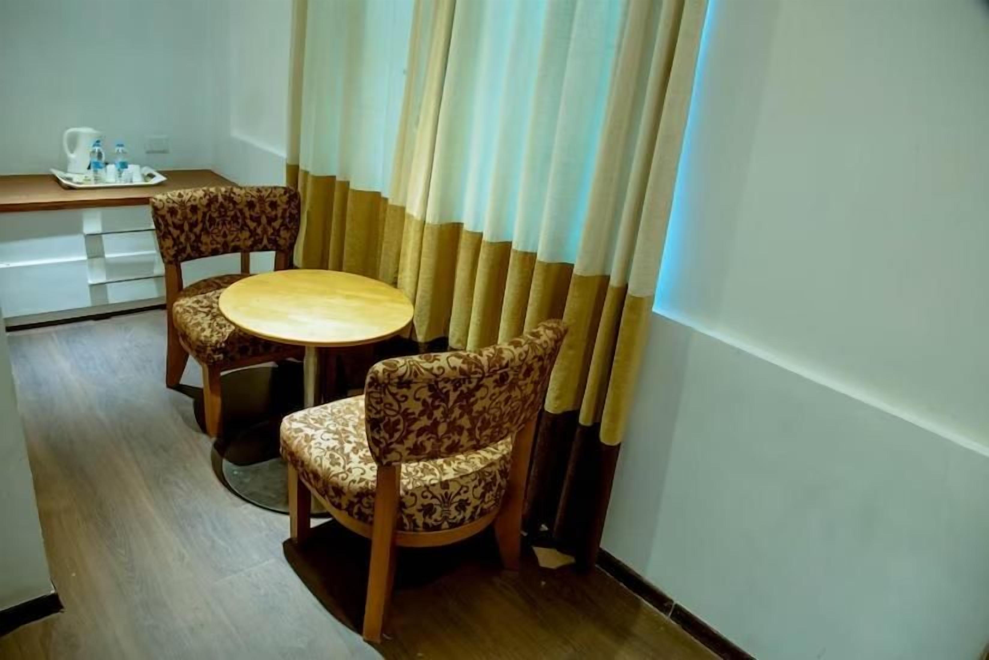 A small dining area with a round table and two patterned chairs, a tray with a teapot and cups, and a window with curtains.