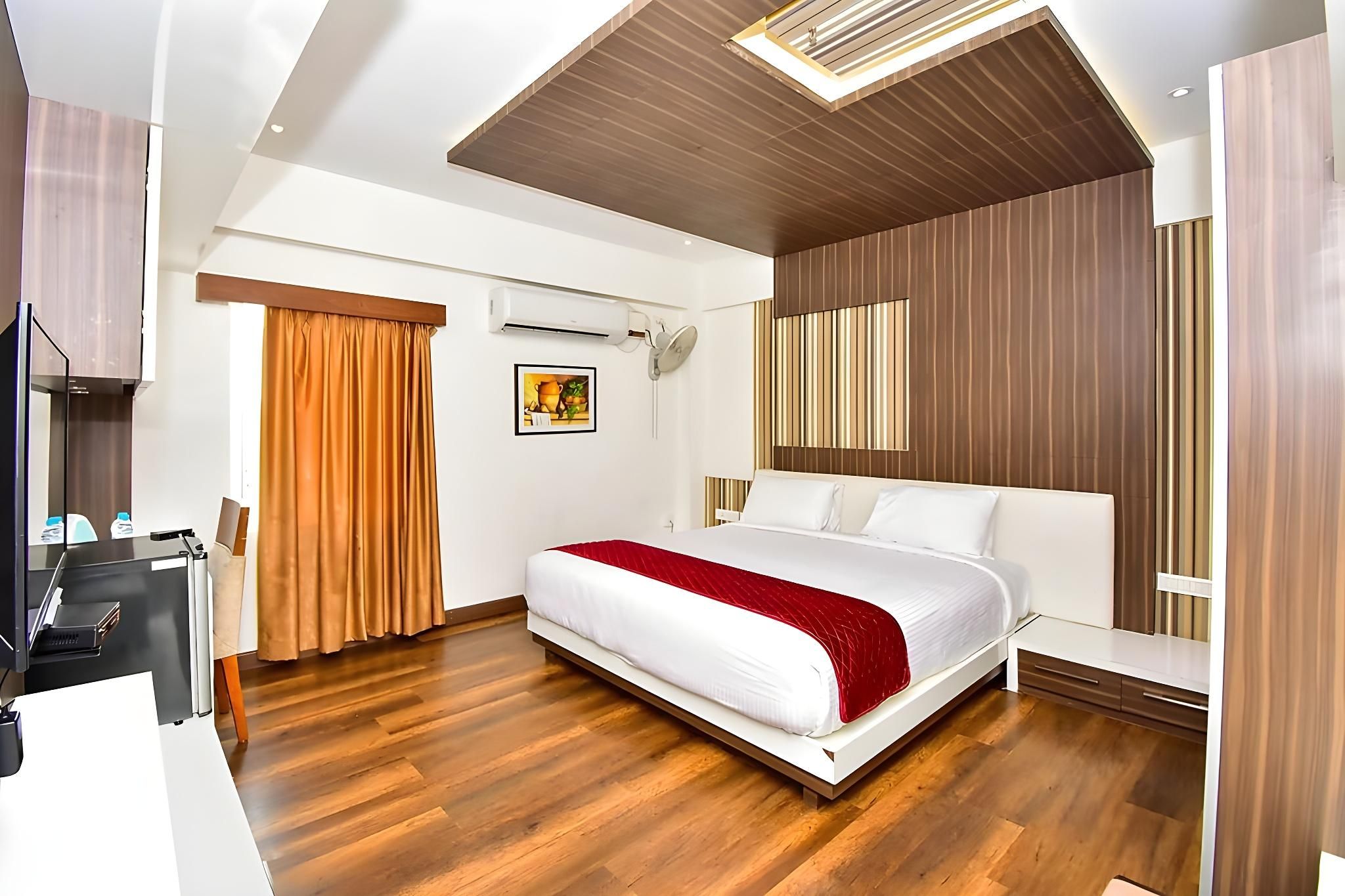 A modern hotel room with a wooden floor, a bed with white and red bedding, a desk with a computer, and an air conditioning unit.