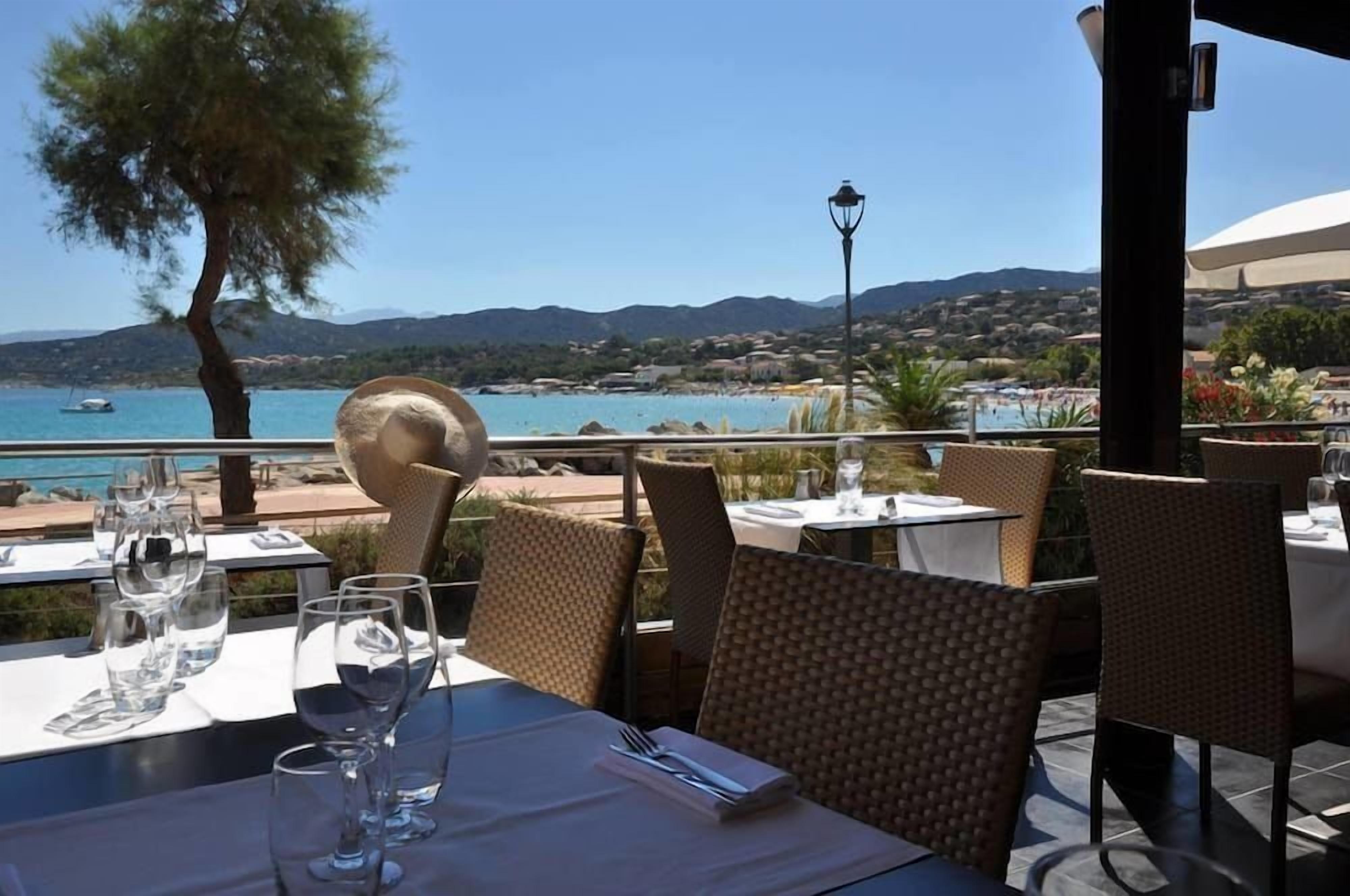 Un espace extérieur aménagé pour les repas, avec des tables et des chaises, offrant une vue sur un paysage côtier sous un ciel dégagé.
