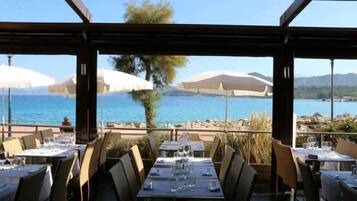 Un espace de restauration en plein air, avec des tables et des chaises, offrant une vue sur un plan d’eau et un paysage montagneux.