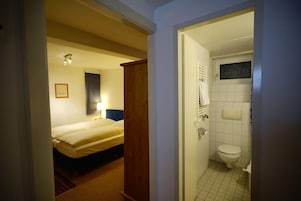Una habitación de hotel con una cama, una lámpara de noche, una puerta de madera y un baño con inodoro y lavamanos.