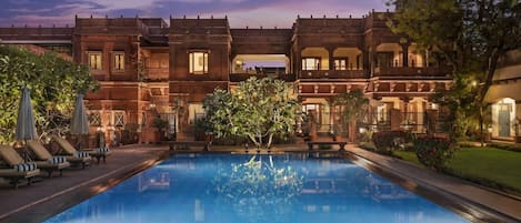 A pool area with a reflection of a building, lounge chairs, and a clear sky.