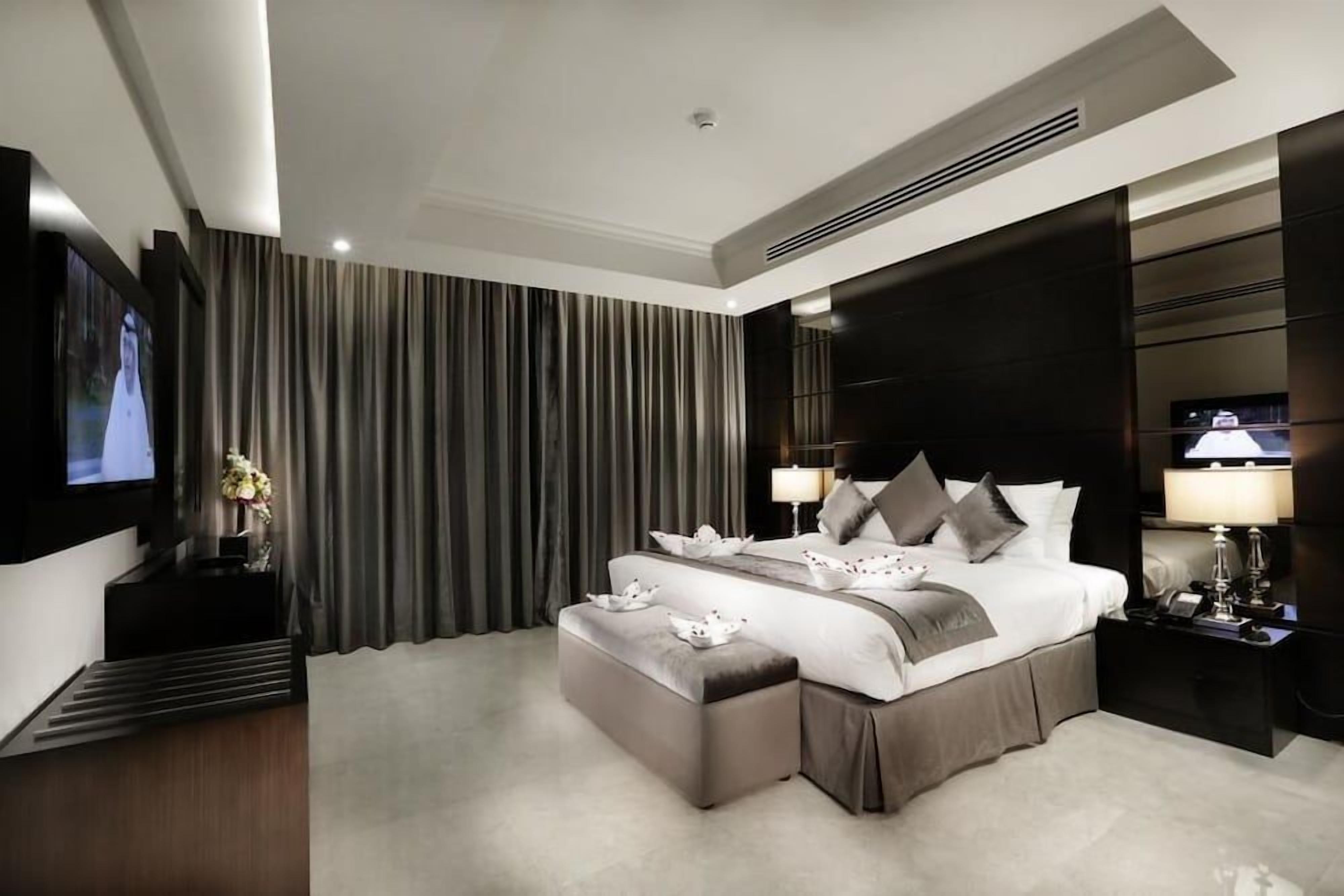 A modern hotel room with a large bed, a flat-screen TV, a bedside table with a lamp, and a large window with curtains.