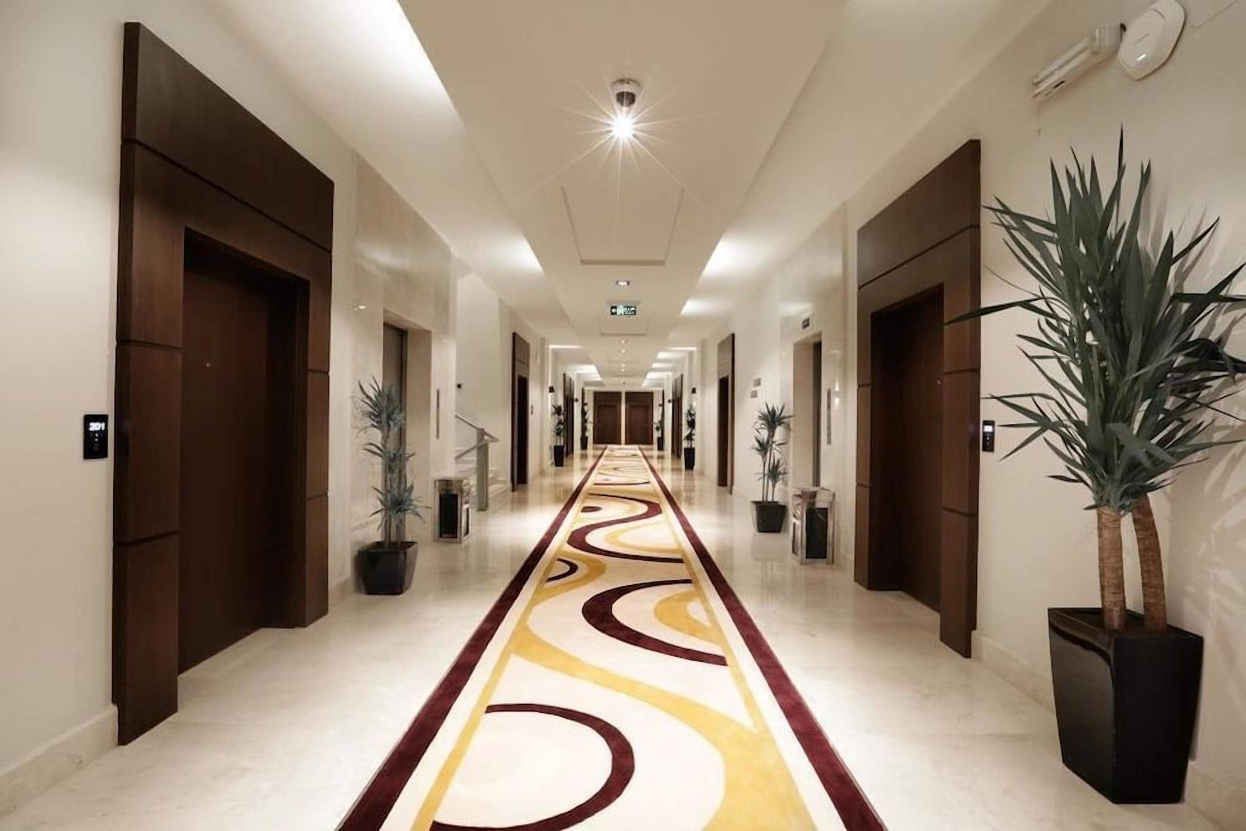 A hotel corridor with a patterned carpet, doors on both sides, and potted plants.