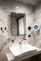 A bathroom with a marble wall, a sink, a mirror, and a wall-mounted light.