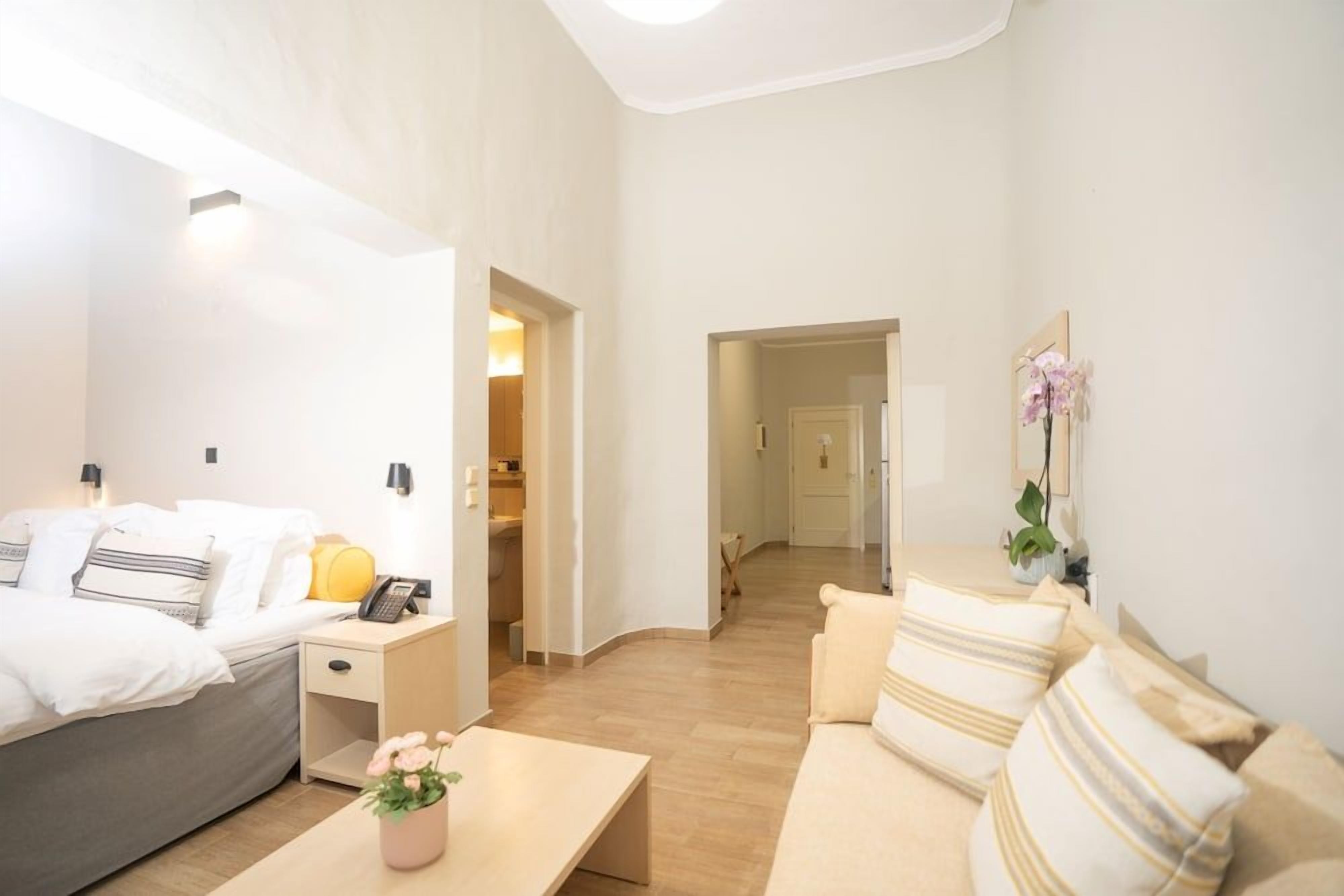 Une chambre d’hôtel avec un lit, une table de chevet, une lampe et une petite table ornée d’une fleur. On y trouve également un canapé et un miroir accroché au mur.