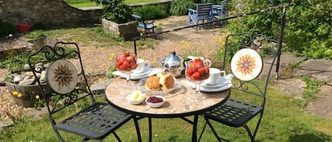 An outdoor dining area with a round table and chairs, surrounded by a garden with a stone wall and greenery.
