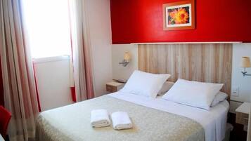 Quarto de hotel com uma cama, duas almofadas, mesa de cabeceira com abajur e janela com cortinas.