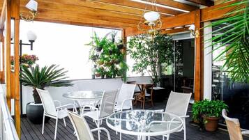 Um pátio externo com móveis de vime branco, mesa de vidro e plantas penduradas.