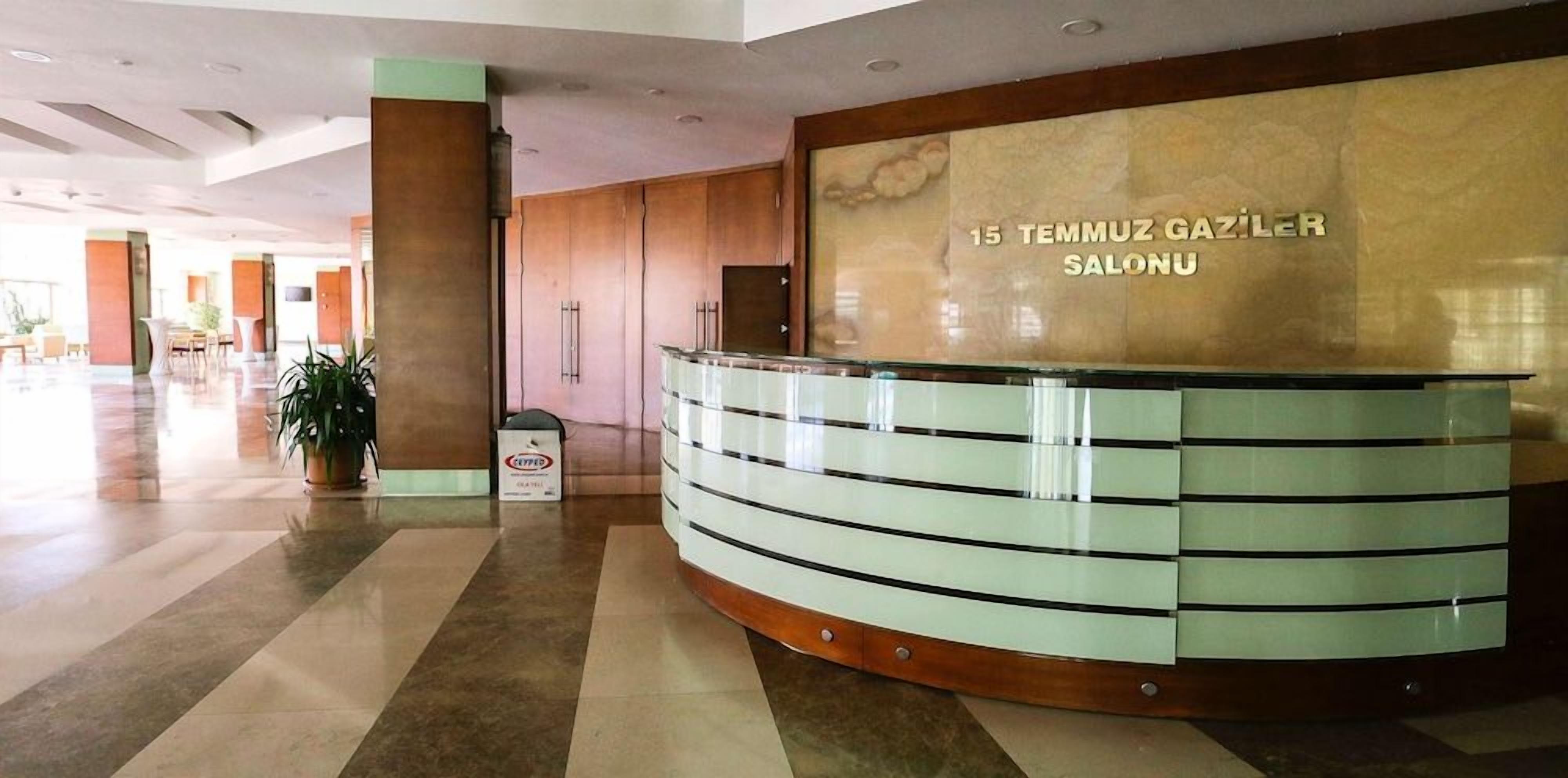 Ein Empfangsschalter mit einem Schild, das '15 Temmuz Gaziler Salonu' zeigt, in einer geräumigen Lobby mit Fliesenboden und einem Blumentopf.
