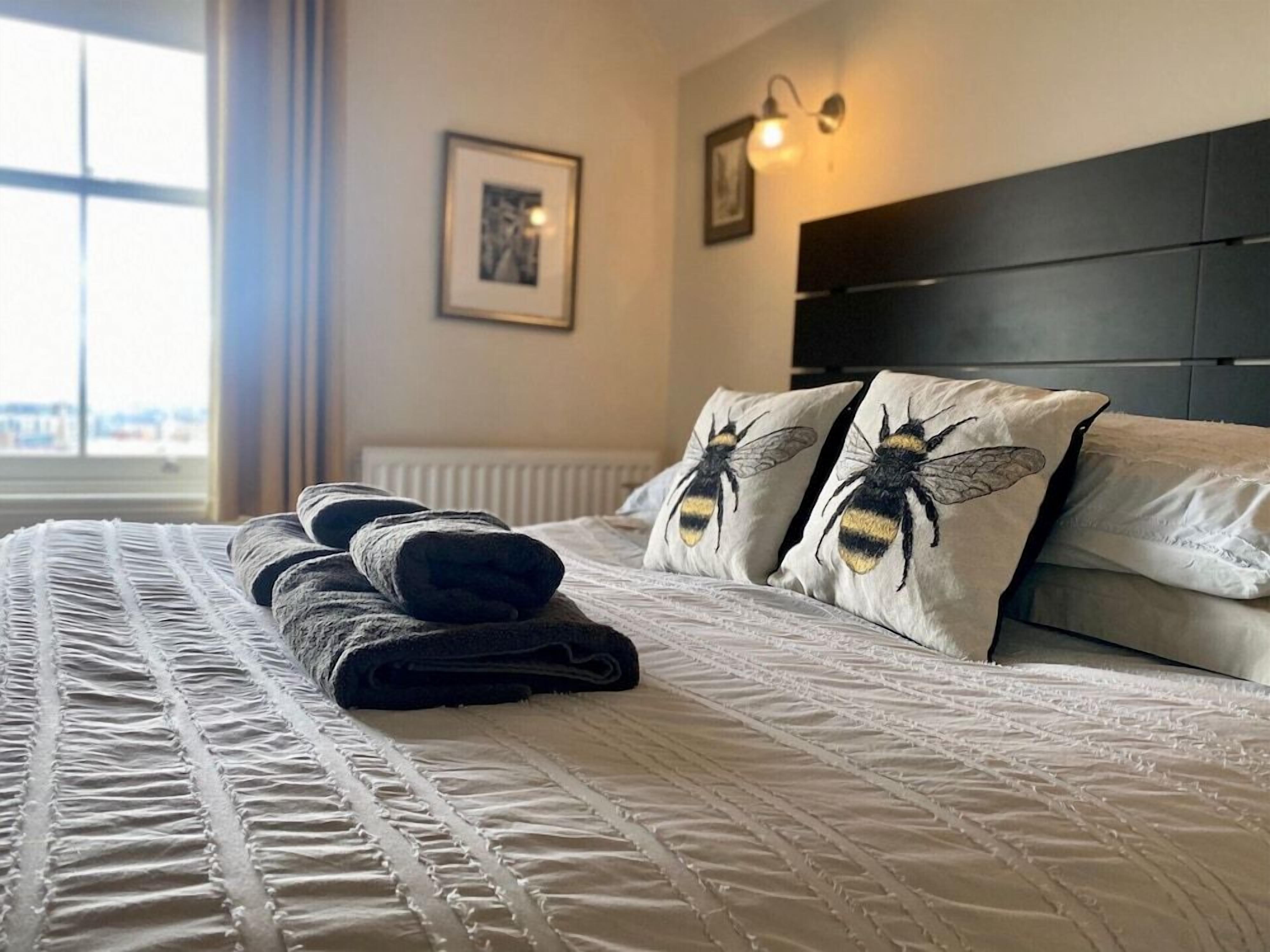 A neatly made bed with a quilted cover, two pillows with a bee design, and a folded towel on the bed.