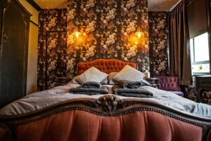 Una cama con cabecera capitonada, un estampado de papel tapiz floral y dos lámparas fijadas a la pared.