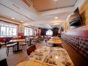 Een restaurantinterieur met meerdere tafels en zitjes, een tafelkleed met patroon en een aan de muur bevestigde televisie.