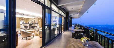 Ein moderner Hotelbalkon mit Meerblick, ausgestattet mit Holztischen und -stühlen, und eine Glaswand, die zu einem Essbereich führt.