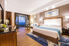 A hotel room with a large bed, a desk, a chair, a small table, and a view of the sea through the balcony doors. - Fengyue Hotel (Huizhou)