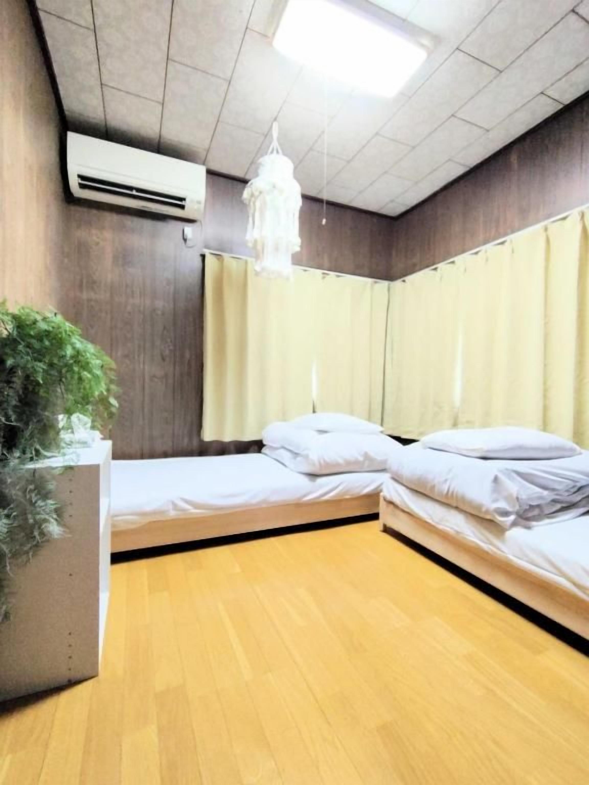 A room with two single beds, a wooden floor, a potted plant, a hanging light fixture, and a ceiling-mounted air conditioning unit.