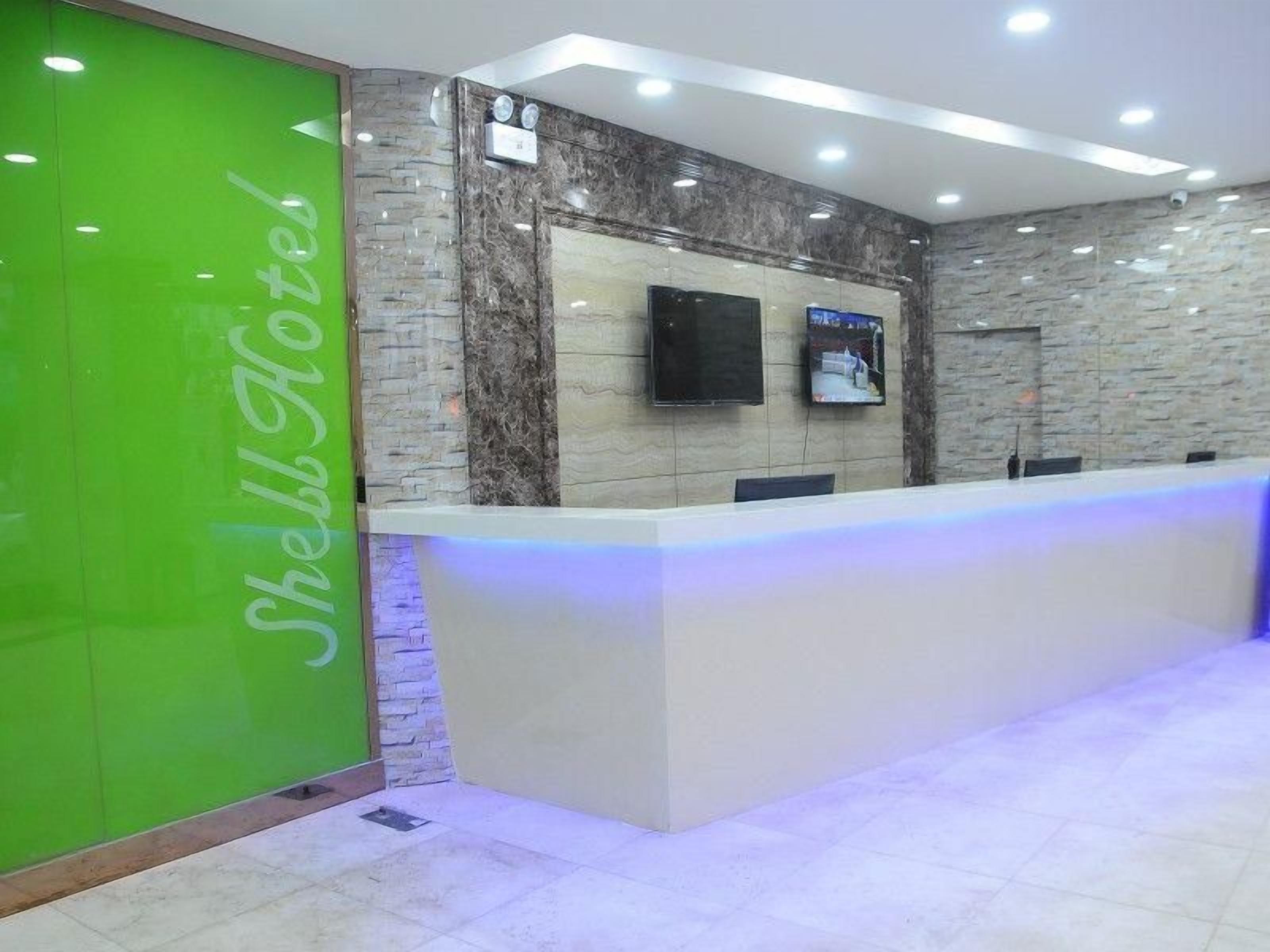 A hotel reception area with a reception desk, a green wall with the hotel's name, a flat-screen TV, and a stone wall.