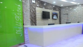 A hotel reception area with a reception desk, a green wall with the hotel's name, a flat-screen TV, and a stone wall.