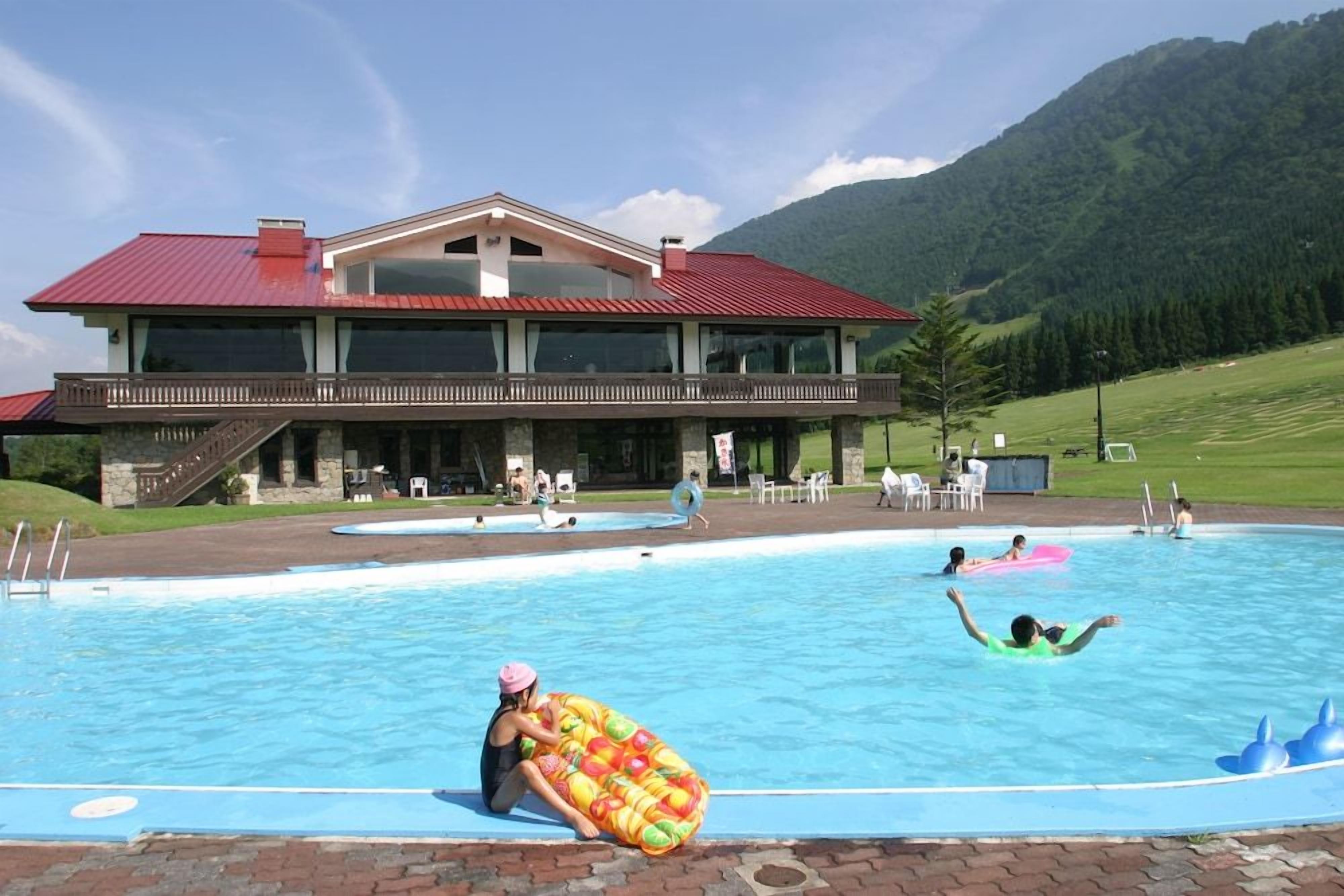 A swimming pool with a building in the background, surrounded by a grassy area and mountains.