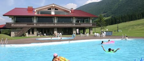Una piscina con un edificio al fondo, rodeada de césped y montañas.