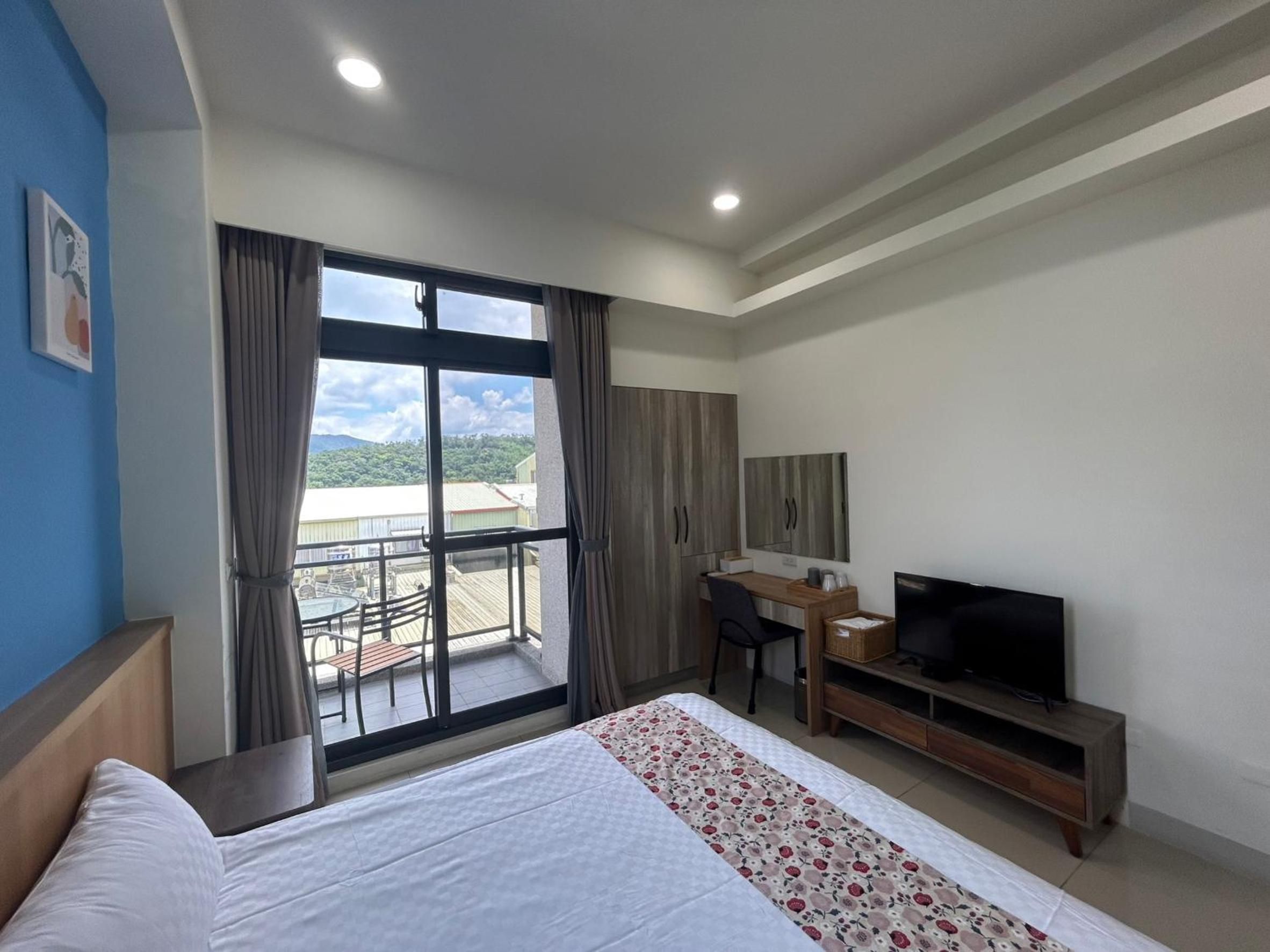 A hotel room with a bed, a desk with a television, and a balcony with a view of the city.