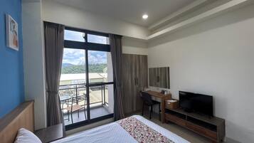 A hotel room with a bed, a desk with a television, and a balcony with a view of the city.