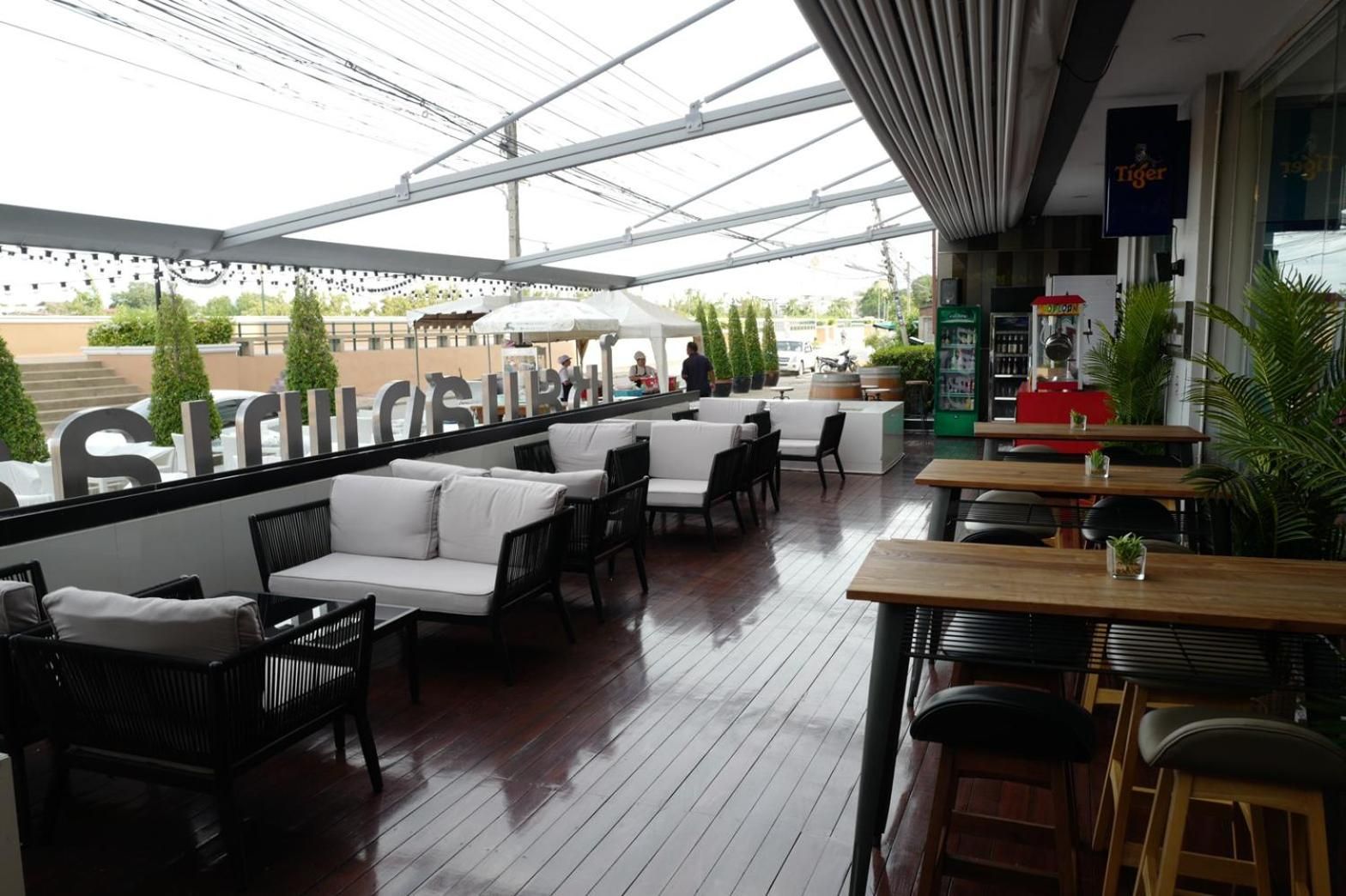 Een terras met witte, beklede banken en houten tafels, onder een glazen dak.
