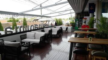 An outdoor seating area with white cushioned sofas and wooden tables, under a glass roof.