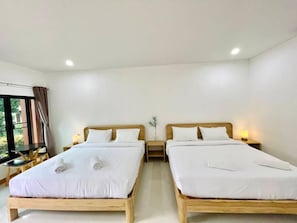 Habitación de hotel con dos camas de madera, ropa blanca, una mesita con una lámpara y una ventana con cortinas.