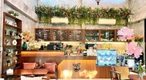 Un café acogedor con mostrador de madera, estantes con plantas y una zona de estar con cojines.