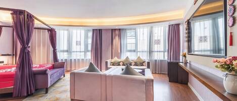 Una habitación de hotel moderna con sofá, cama con cortinas moradas y un ventanal con cortinas de gasa.