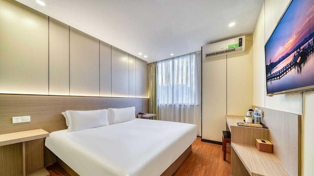 Ein Hotelzimmer mit einem großen Bett, einem Nachttisch, einem Schreibtisch, einem Sessel, einer Klimaanlage und einem an der Wand befestigten Fernseher.