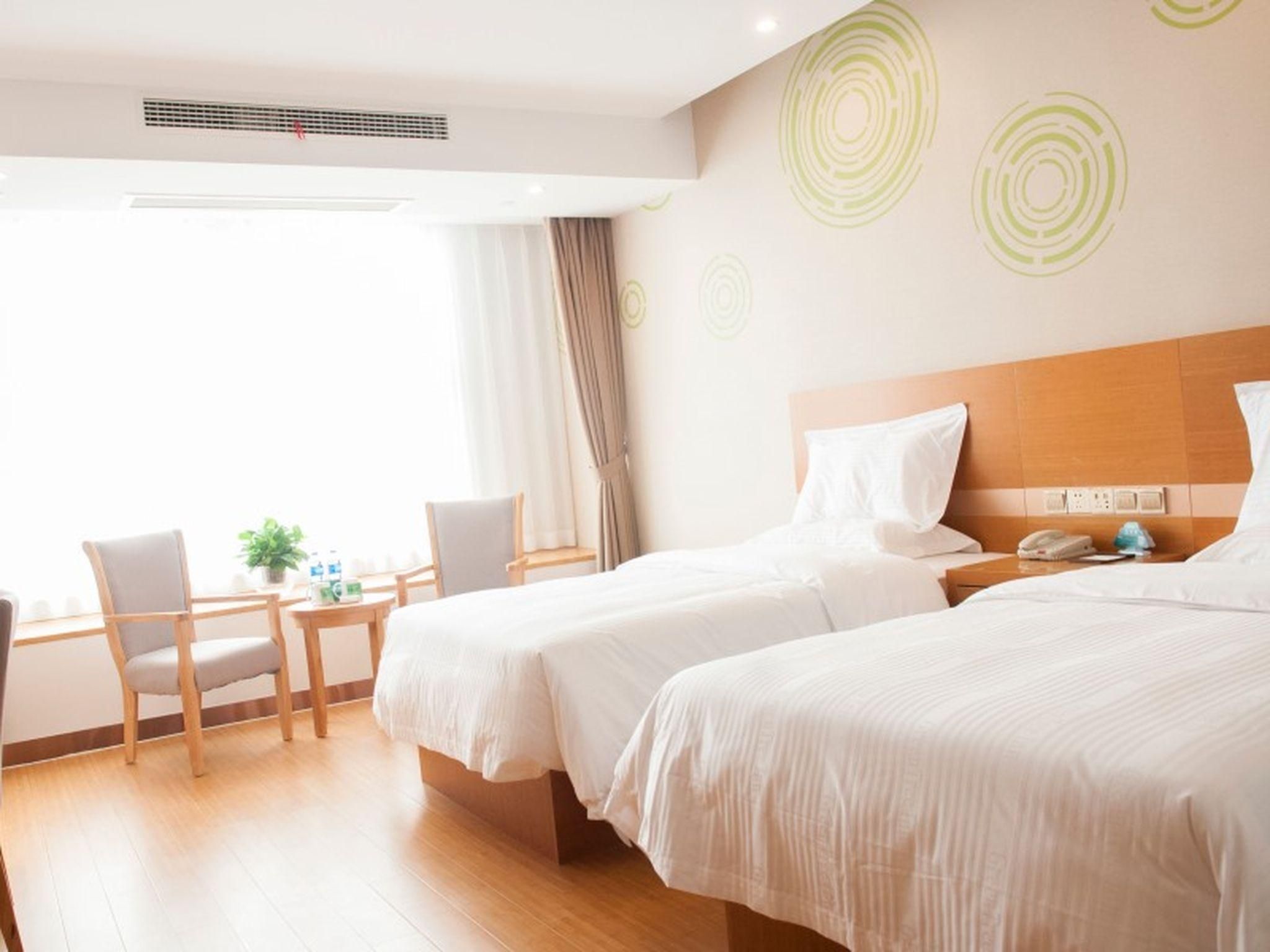 A hotel room with two beds, a desk with chairs, a small table with a plant, and a window with curtains.