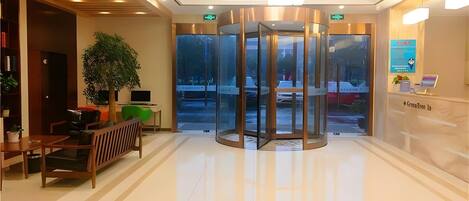 A modern lobby with a revolving door, a reception desk, and a seating area with a plant and a laptop.