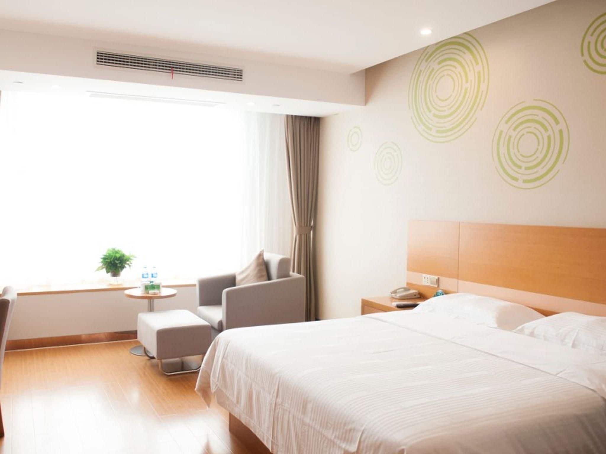 Een hotelkamer met een groot bed, een nachtkastje met een lamp, een stoel, een klein tafeltje met een plant en een raam met gordijnen.