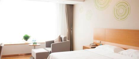 Een hotelkamer met een groot bed, een nachtkastje met een lamp, een stoel, een klein tafeltje met een plant en een raam met gordijnen.