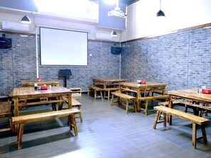 Un salón de conferencias con varias mesas y bancos de madera, una pantalla de proyector y una pared de ladrillo.