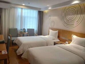 A hotel room with two beds, a desk, a chair, and a small table with a lamp.