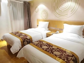 A hotel room with two beds, each with a white bedspread and a decorative gold trim. There is a nightstand with a lamp and a phone on it.