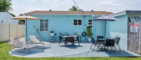 A backyard with a blue house, a patio with chairs and tables, a bicycle, and a notice sign.