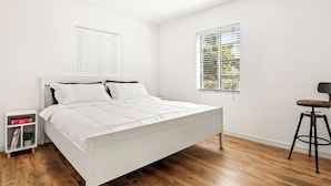 Un dormitorio con cama blanca, suelo de madera, mesita de noche, ventana con persianas y una silla.